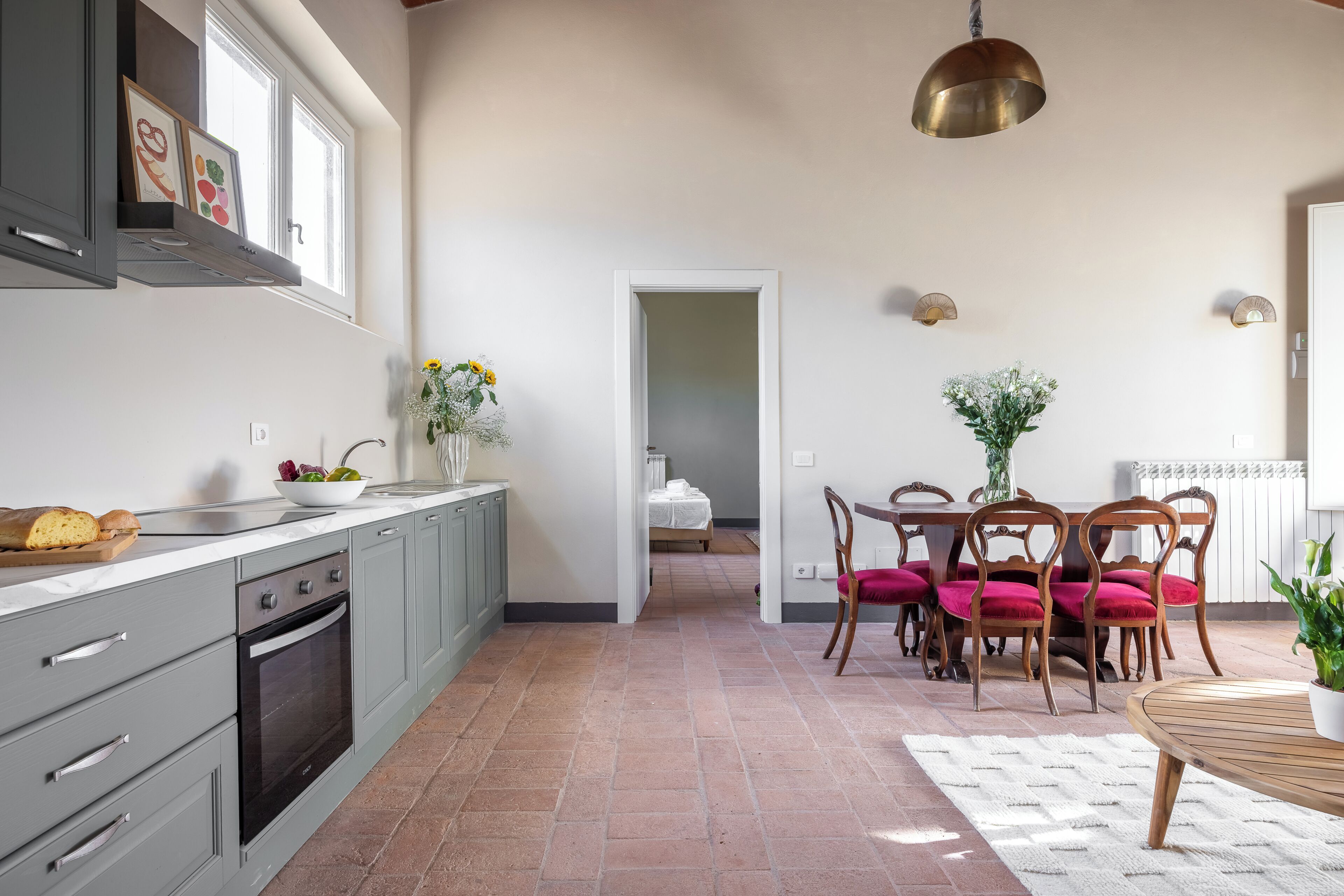 A kitchen with grey cabinets, a wooden dining table with red chairs, and a hanging brass light fixture.