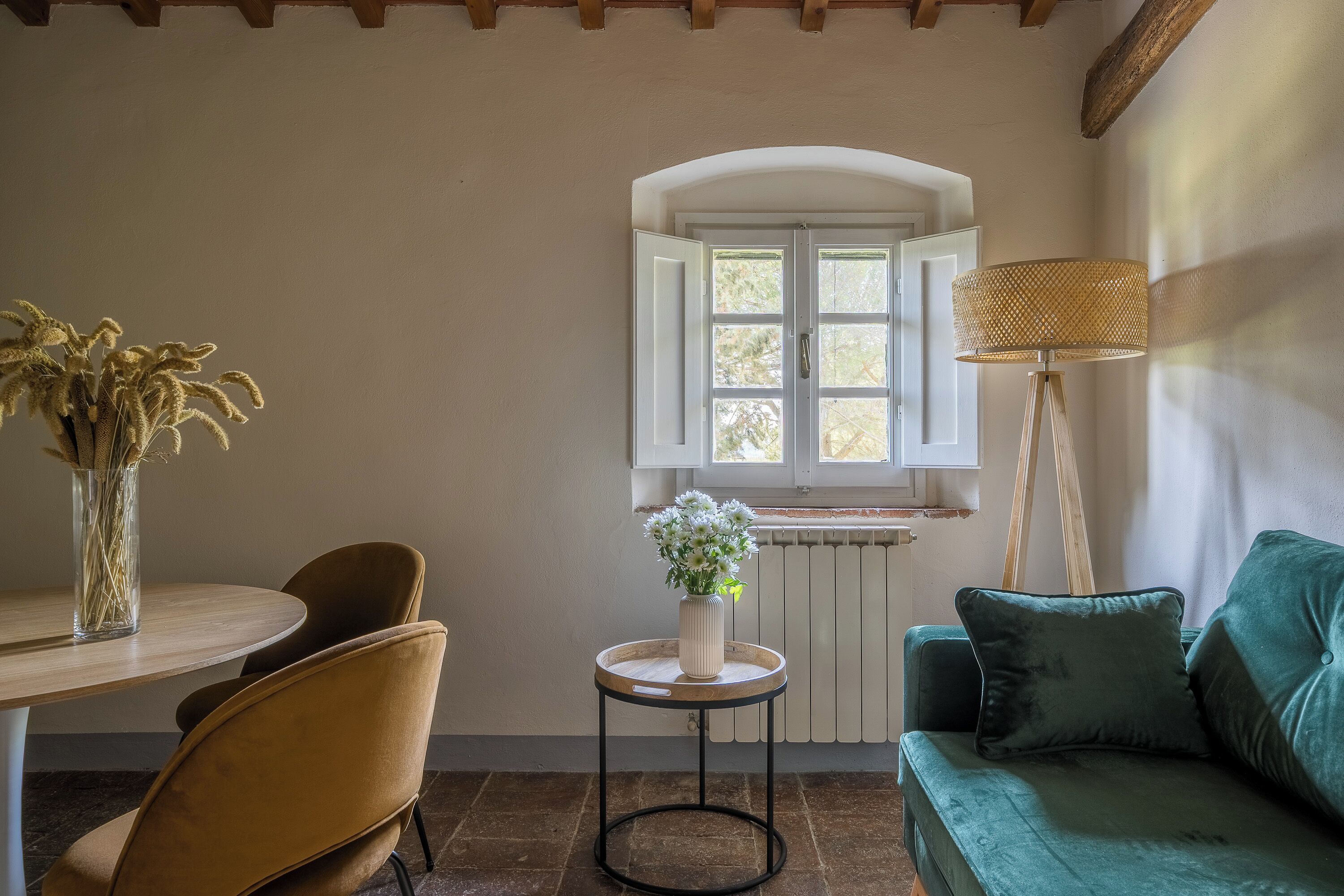 Una habitación acogedora con una mesa redonda de madera, un jarrón con flores secas, un sofá verde y una lámpara.