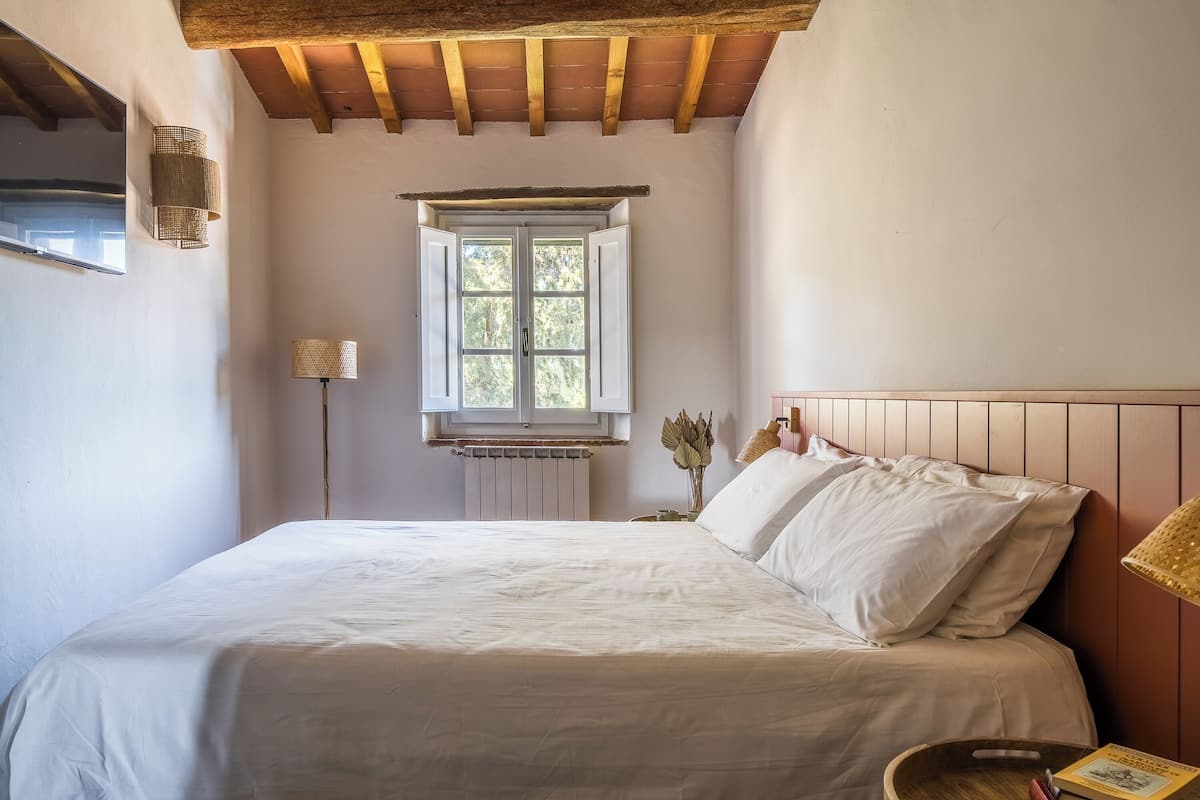 Un dormitorio con una cama grande, vigas de madera en el techo, una ventana con persianas blancas y una mesita de noche con una lámpara y un libro.