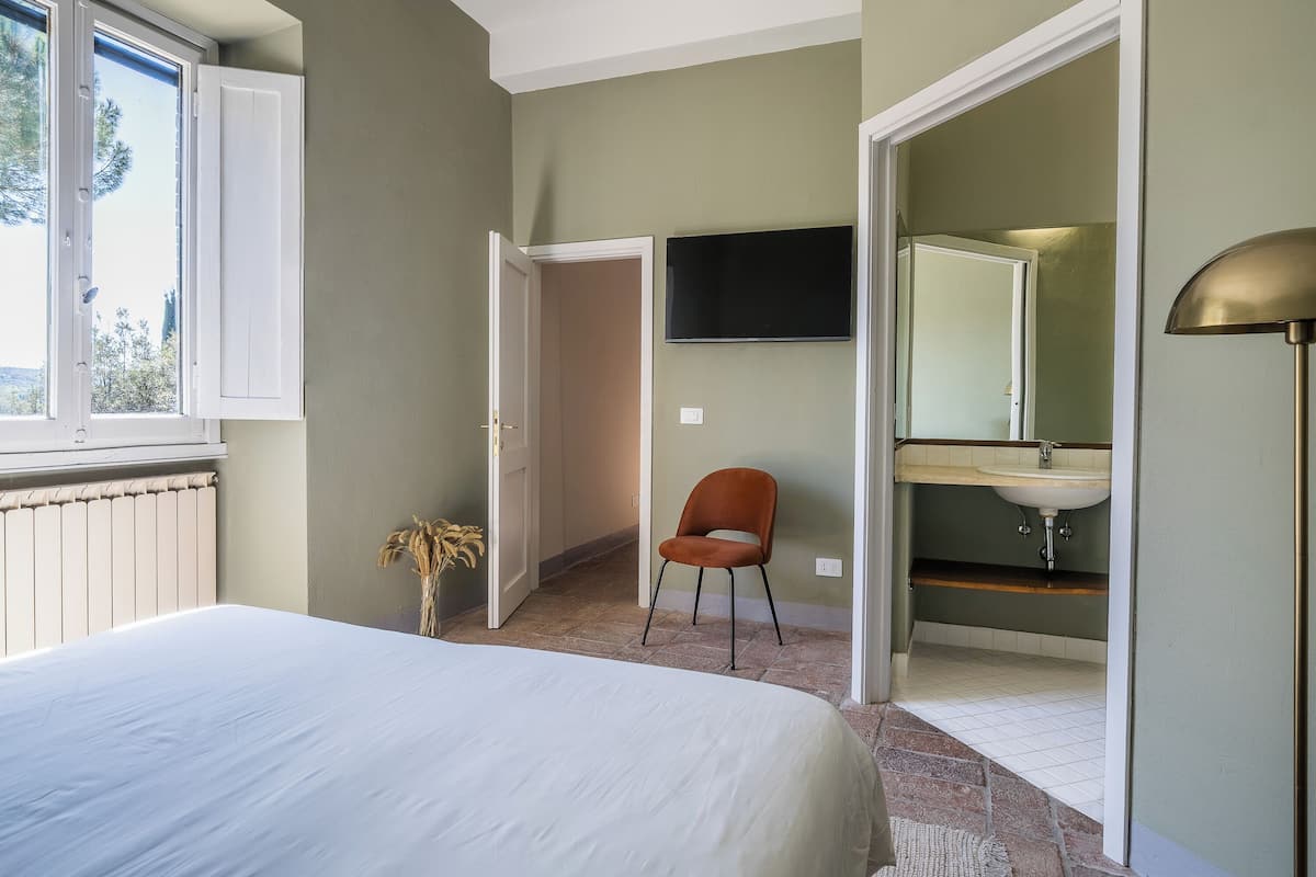 Un dormitorio con cama, una silla, televisor y baño con lavamanos y espejo.