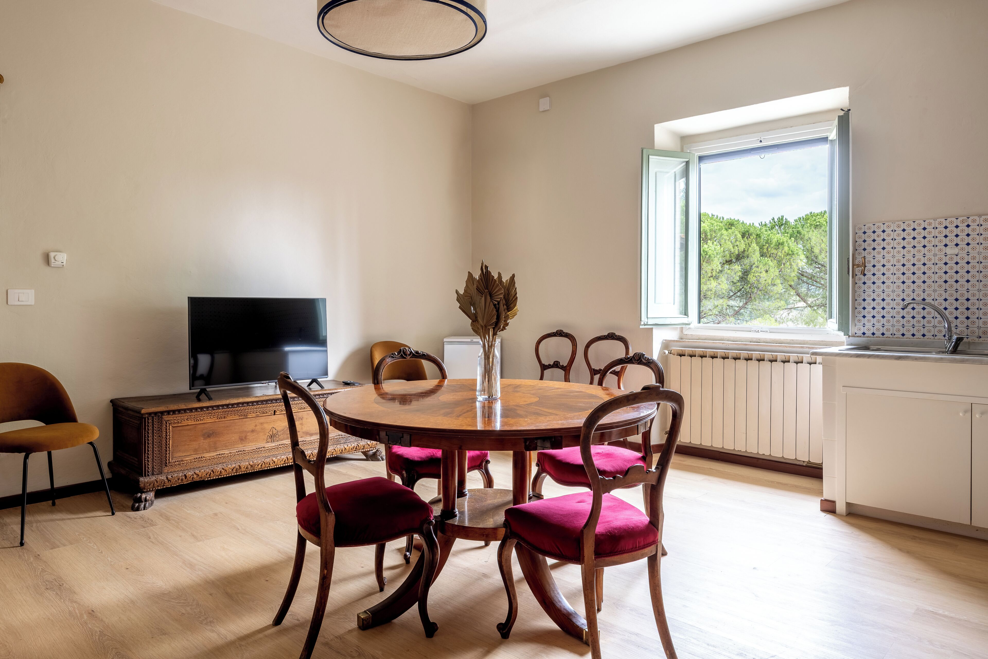 A dining area with a round wooden table, six chairs, a vase with dried flowers, a television on a wooden cabinet, and a window with a view of trees.