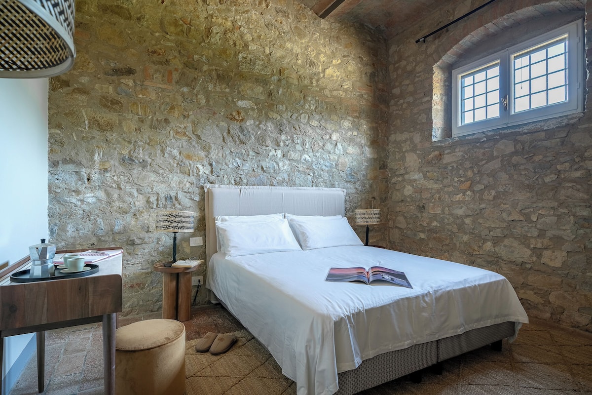 Un dormitorio con paredes de piedra, una cama, mesita de noche, lámpara y ventana.