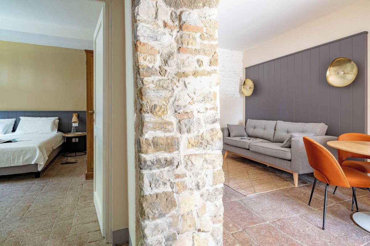 Habitación de hotel con pared de piedra, una cama, un sofá y un área de comedor con sillas naranjas.