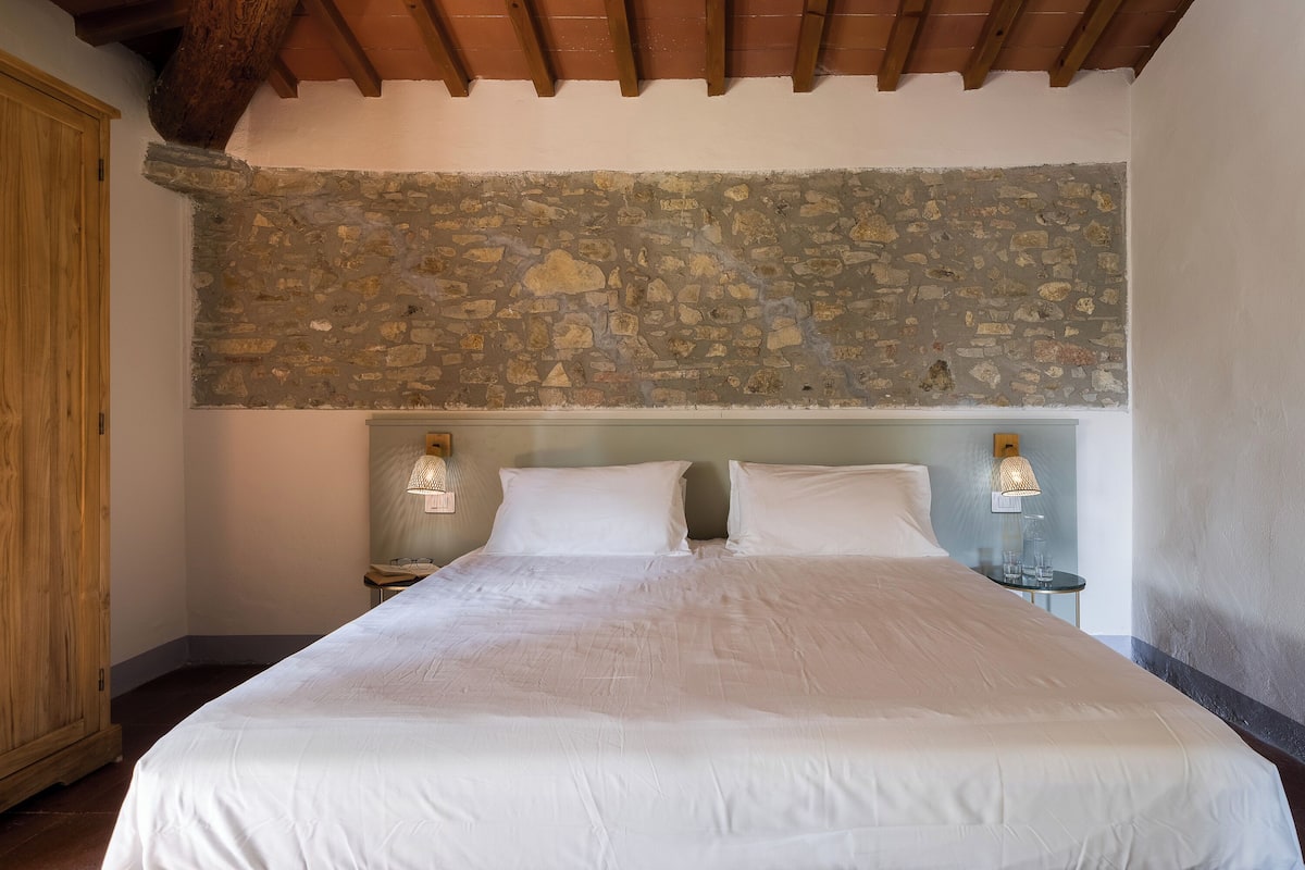 Un dormitorio con pared de piedra, vigas de madera, una cama con ropa blanca, dos lámparas de noche y un armario de madera.