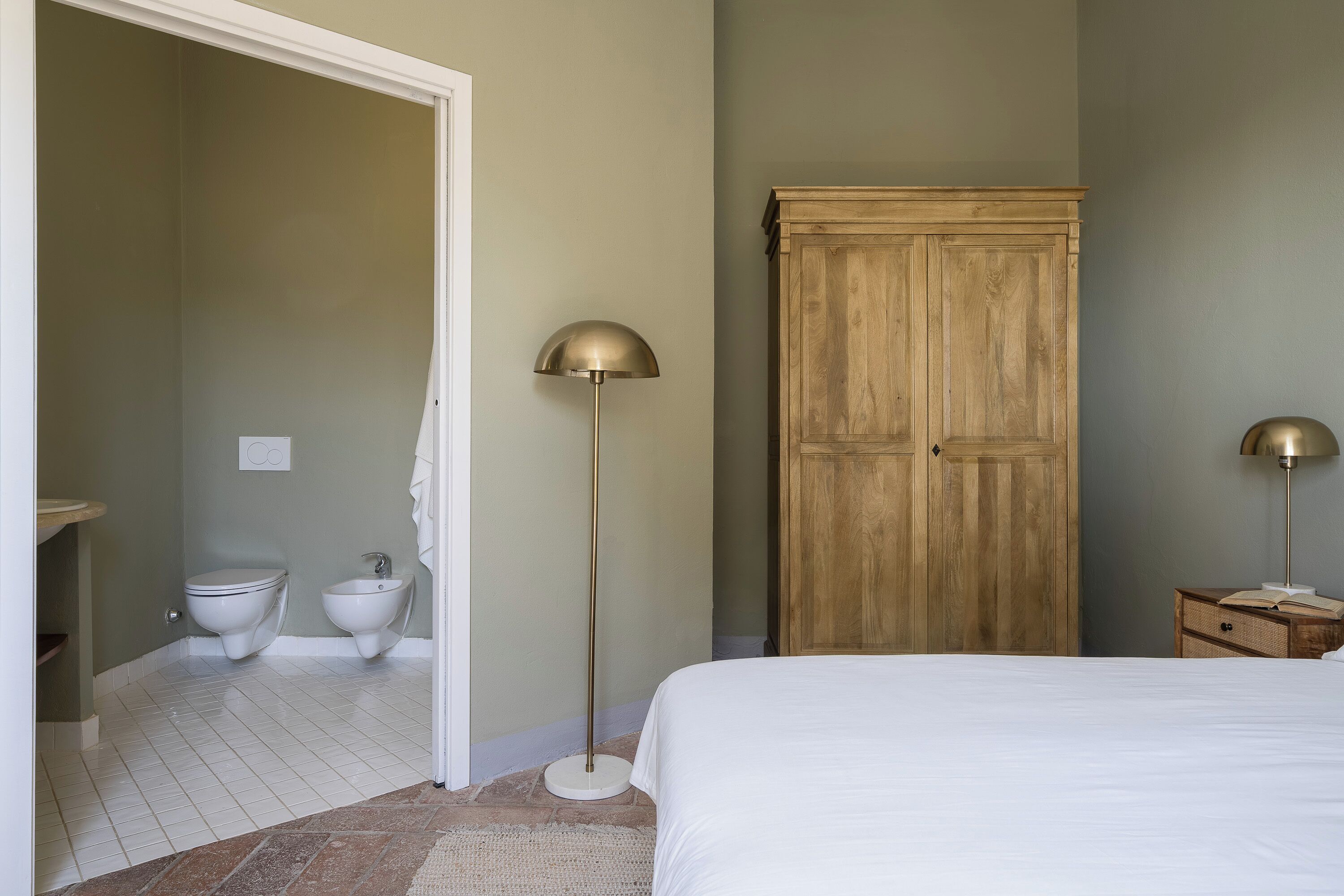 A bedroom with a bed, a wardrobe, a floor lamp, a bedside table with a book, and a bathroom with a toilet and bidet.