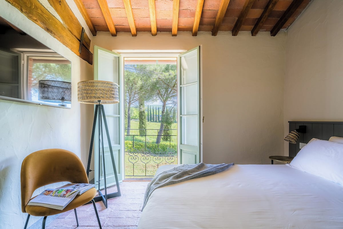 Un dormitorio con cama, una silla con una revista, una lámpara de pie y vistas a través de una puerta abierta.