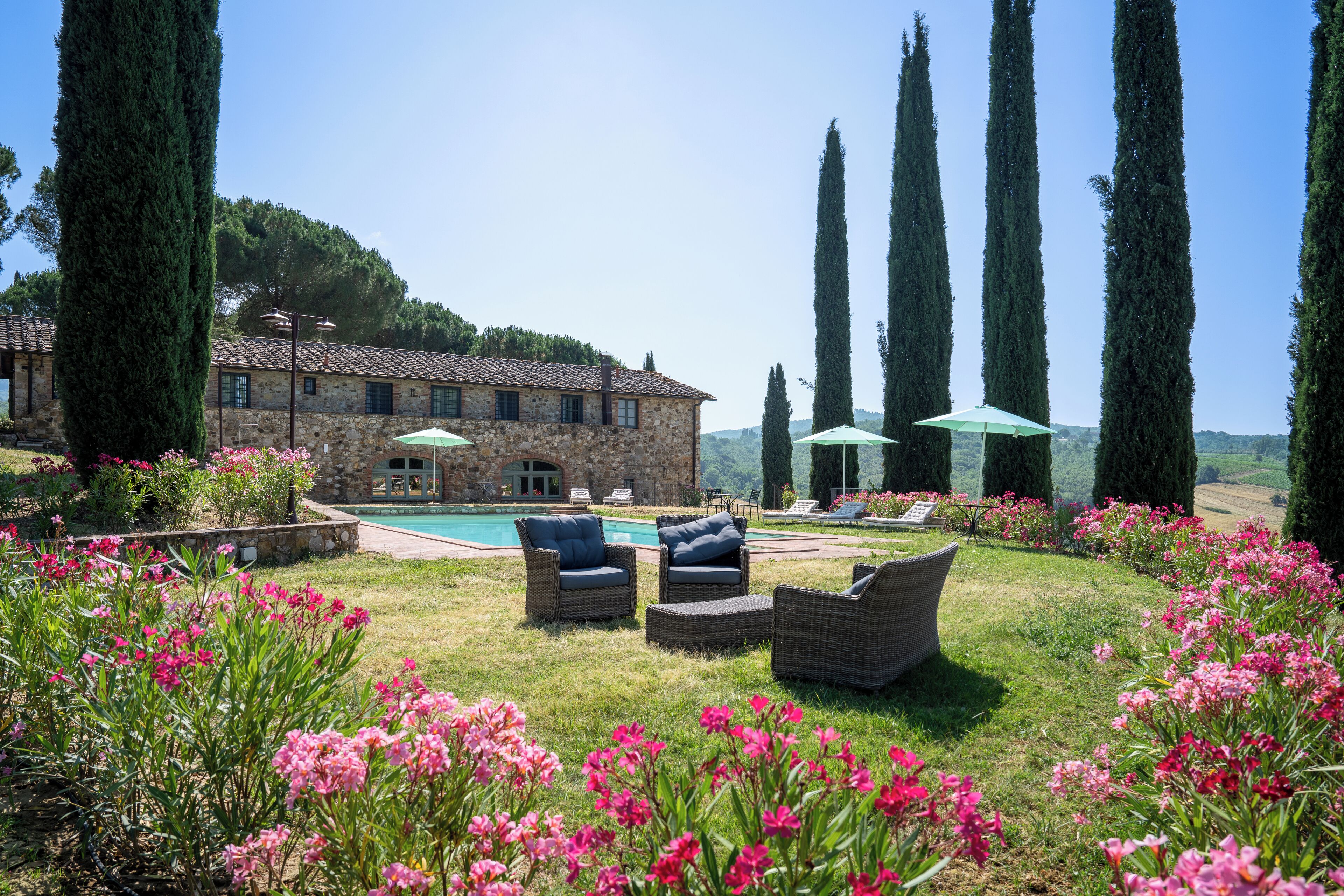 A stone building with a swimming pool, surrounded by tall cypress trees and pink flowers.