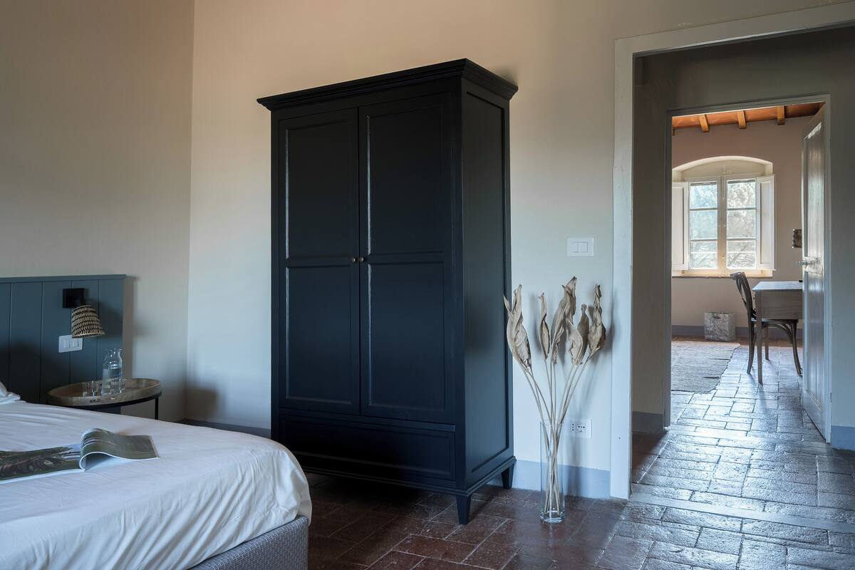 Una habitación de hotel con una cama, un armario de madera oscura, una mesita redonda con una lámpara y un florero con plantas secas.
