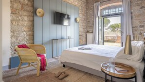 A bedroom with a stone wall, a bed with a white bedspread, a television mounted on the wall, and a window with curtains.