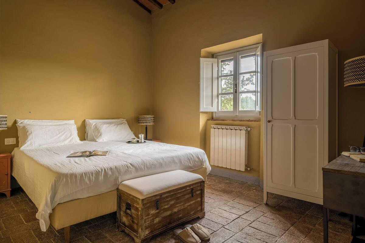 Un dormitorio con cama, mesita de noche, lámpara, armario y ventana con vista a árboles.