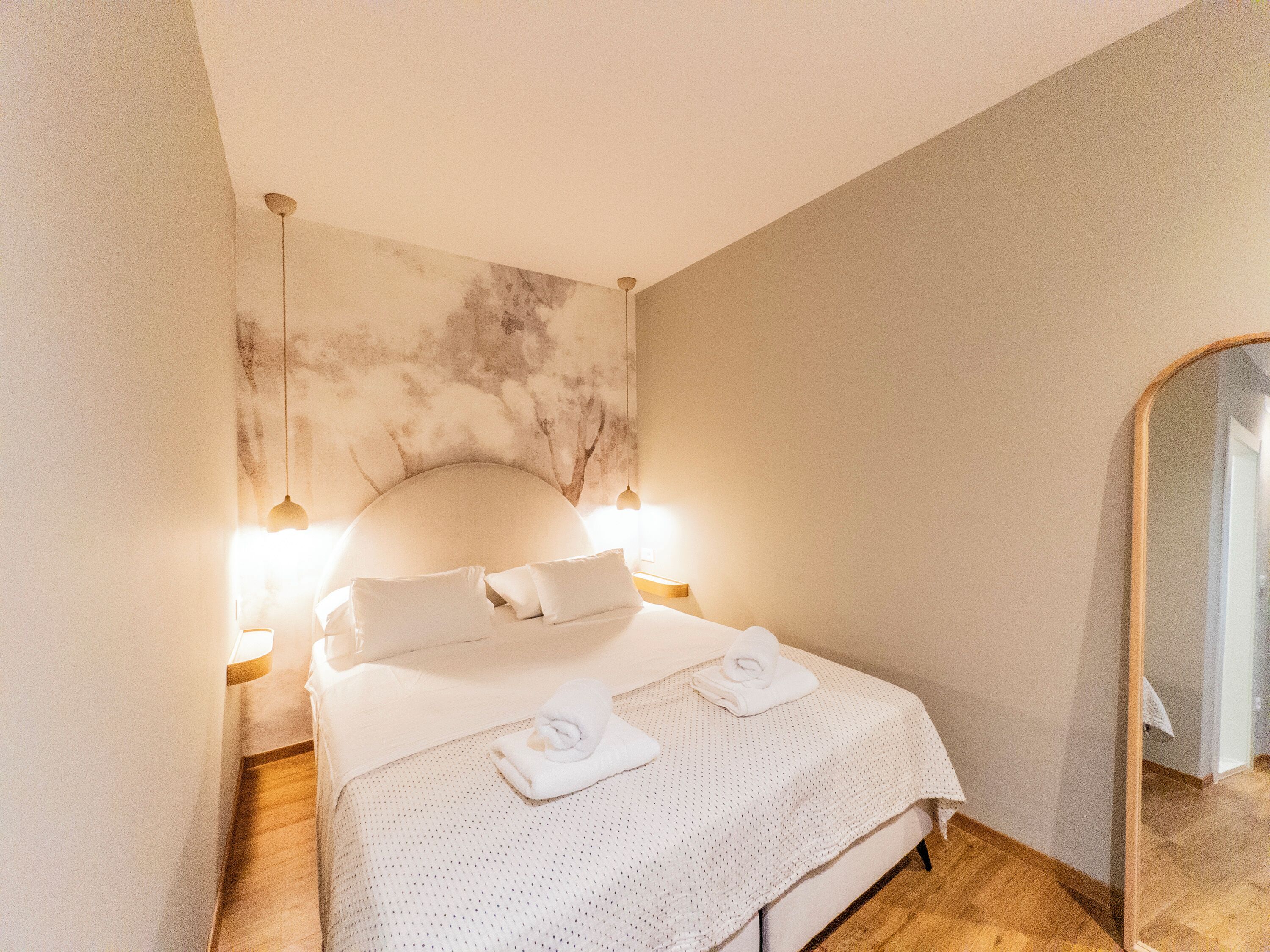 A neatly made bed with white linens and two towels, a wall-mounted headboard, and a large mirror on the right.