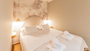 Cama bem arrumada com lençóis e almofadas brancas, duas luminárias de cabeceira e parede com textura decorativa.
