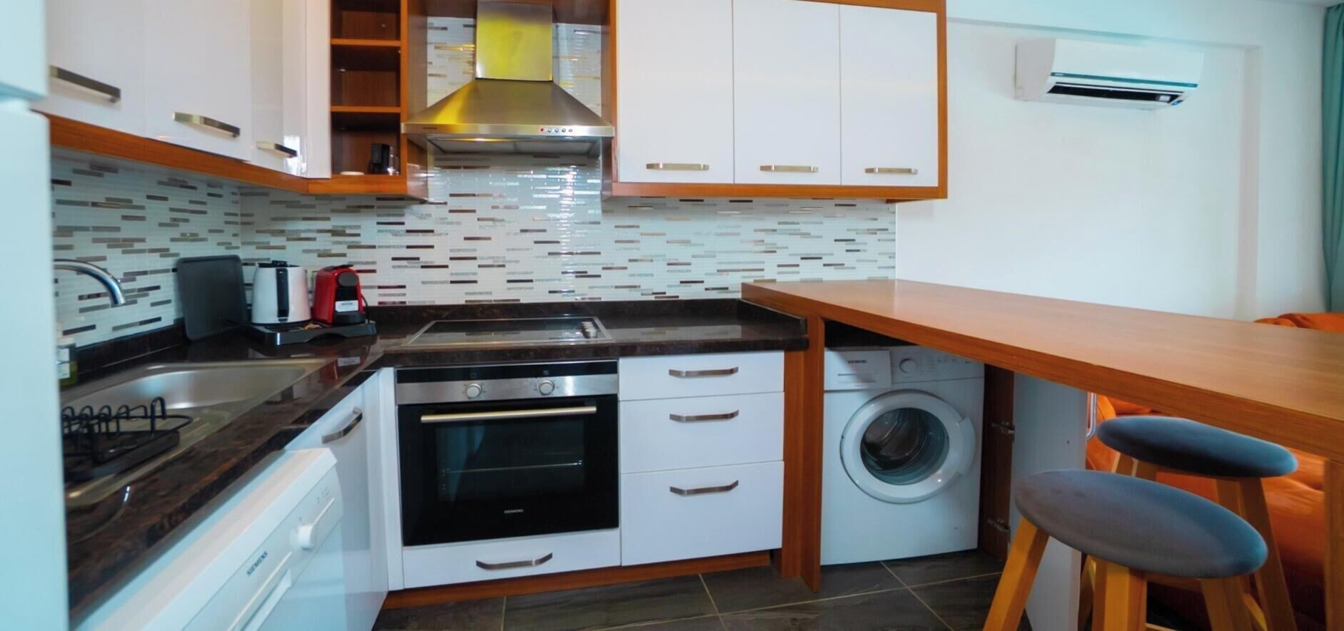 A compact kitchen with a stove, oven, washing machine, and a wooden dining area.