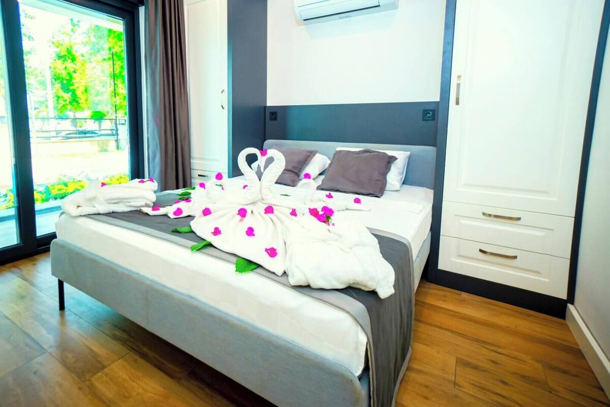 A modern hotel room with a bed, a window with a view of greenery, a built-in air conditioning unit, and a wooden floor.