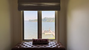 Een hoekzaal met een groot raam met uitzicht op een meer, een bankje en een aanblik van een steiger en een huis aan de overkant van het water.