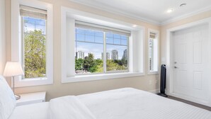 A hotel room with a large bed, a bedside table with a lamp, a TV stand, and a view of the city through large windows.