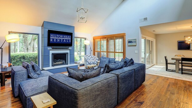 Una sala de estar moderna con un sofá seccional grande, una chimenea y un televisor que muestra 'Hilton Head Rentals'.