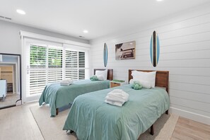 A hotel room with two beds, a large window with blinds, a surfboard on the wall, and a beach-themed painting.