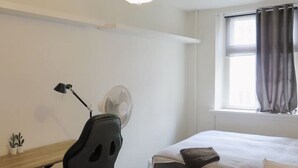 A modern bedroom with a bed, a desk with a chair, a standing fan, a small plant, and a window with curtains. - Close to Norreport st and walking street. 305 (København)