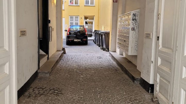 A narrow hallway with a cobblestone floor leading to a yellow building with a parked car. - Close to Norreport st and walking street. 305 (København)