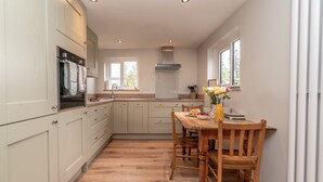A modern kitchen with light-colored cabinets, a wooden dining table, and a window providing natural light. - A Cosy Farm Retreat for Two | Muriau-Bach (Criccieth)
