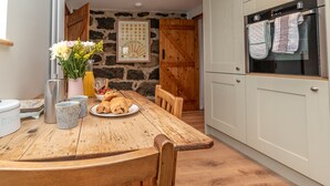 A rustic kitchen with a wooden table, a stone wall, and a modern oven. - A Cosy Farm Retreat for Two | Muriau-Bach (Criccieth)