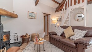 A cozy living room with a brown leather sofa, a wooden staircase, a fireplace, and a wicker basket. - A Cosy Farm Retreat for Two | Muriau-Bach (Criccieth)