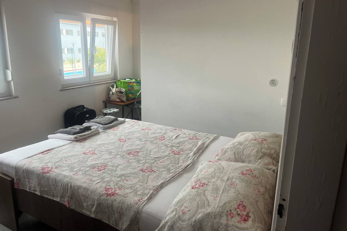 A neatly made bed with floral bedding, a small table with a laptop and bags, and a window with a view of buildings.