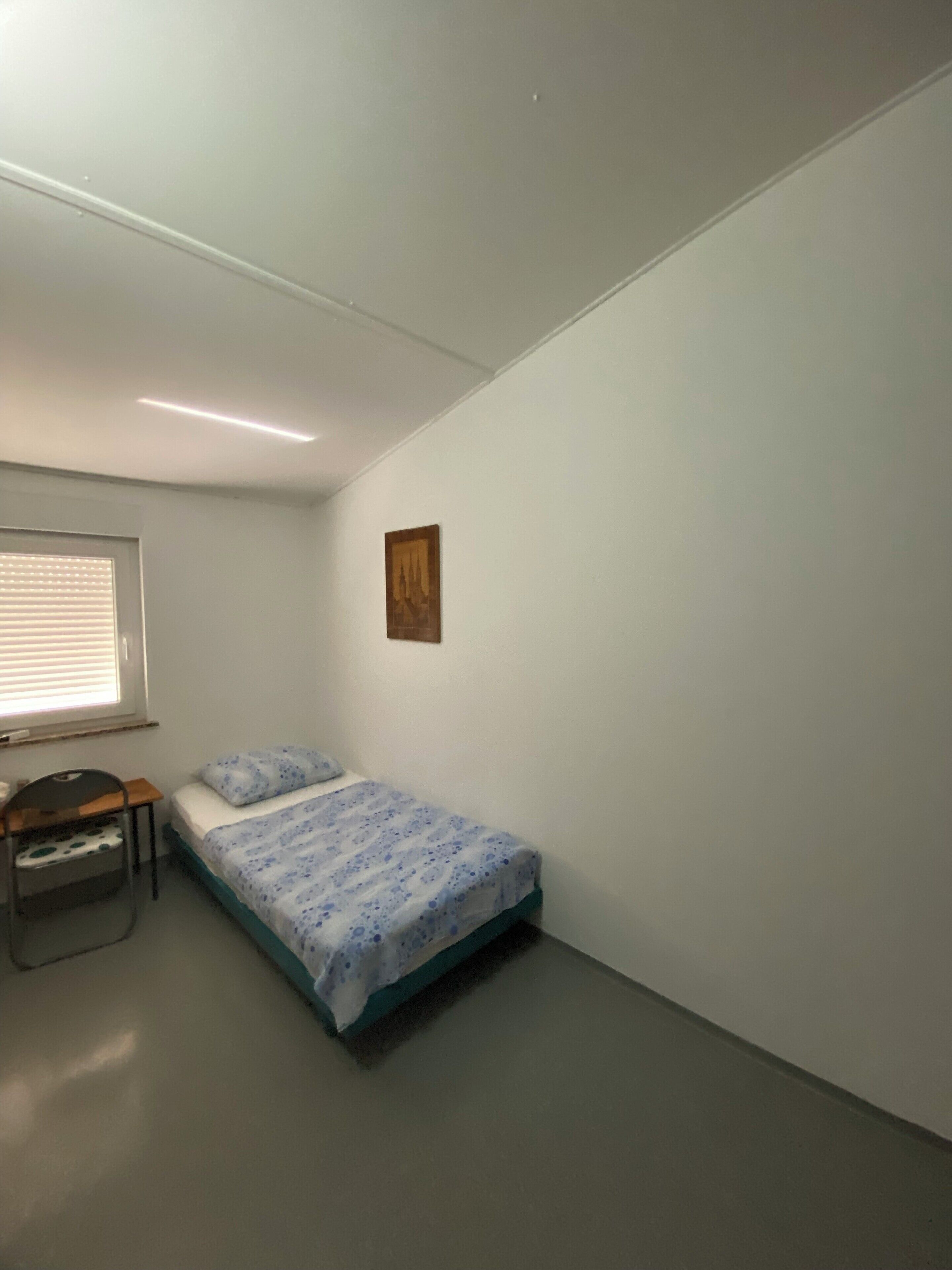 Een kleine, eenvoudig ingerichte kamer met een enkel bed, een bureau en een raam met jaloezieën.
