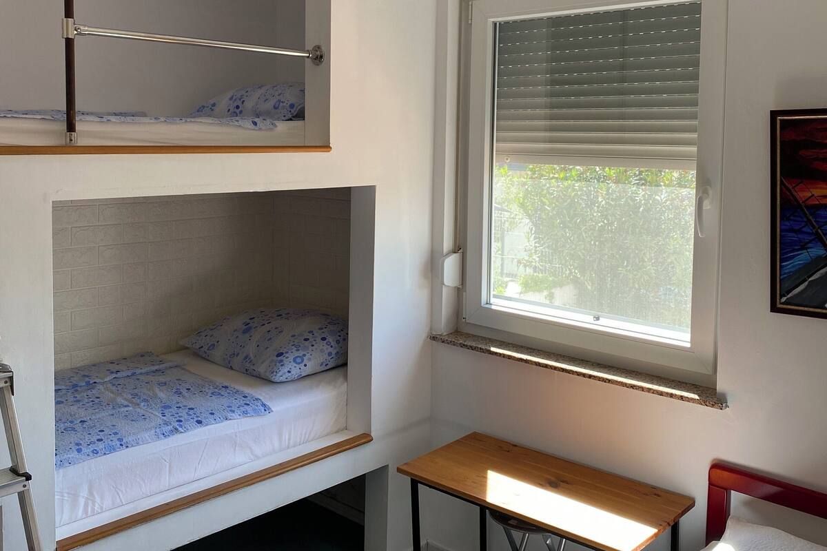 A dormitory room with bunk beds, a window with blinds, a desk, and a ladder.