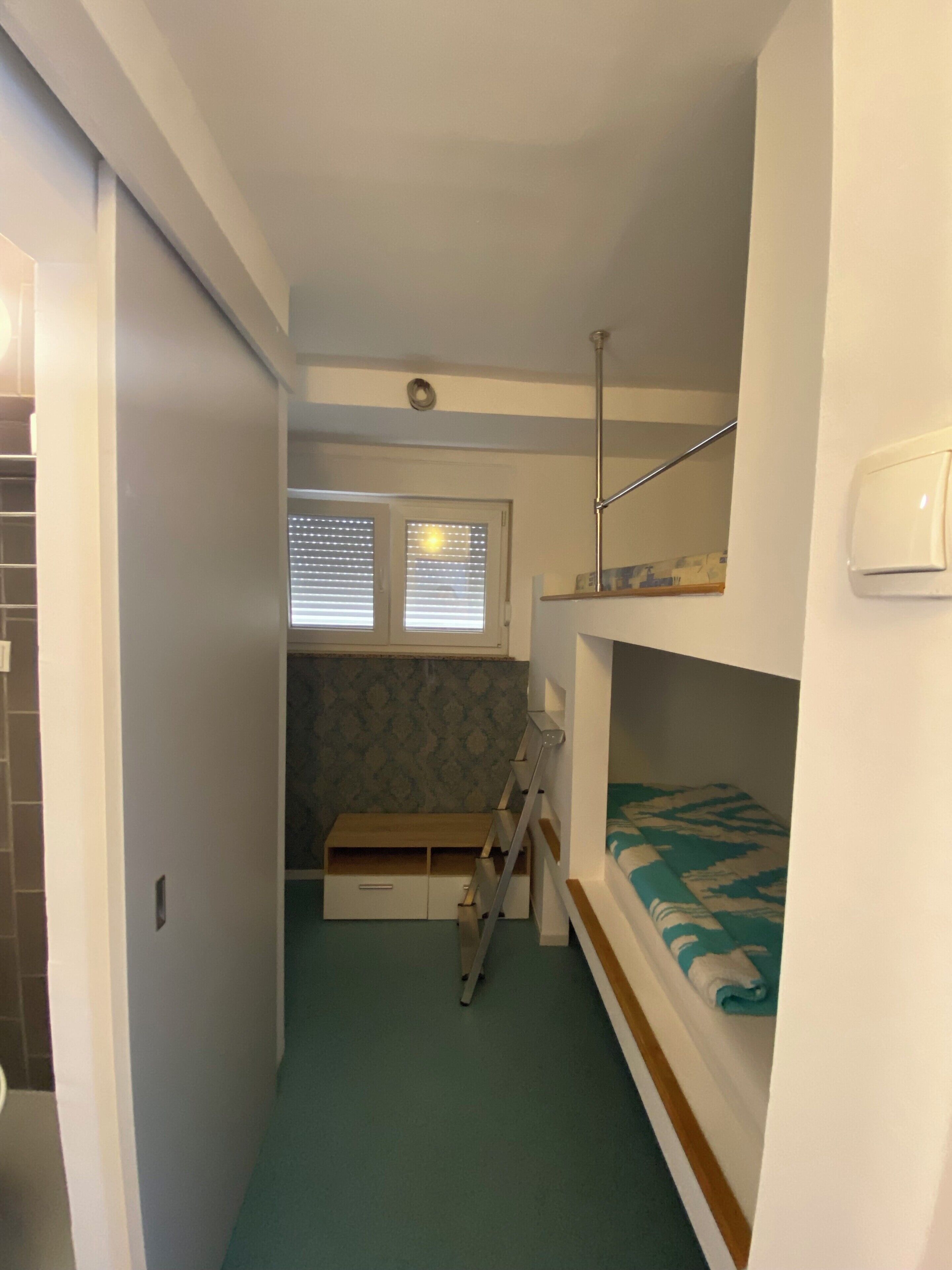 A narrow room with bunk beds, a small desk, and a window with blinds.