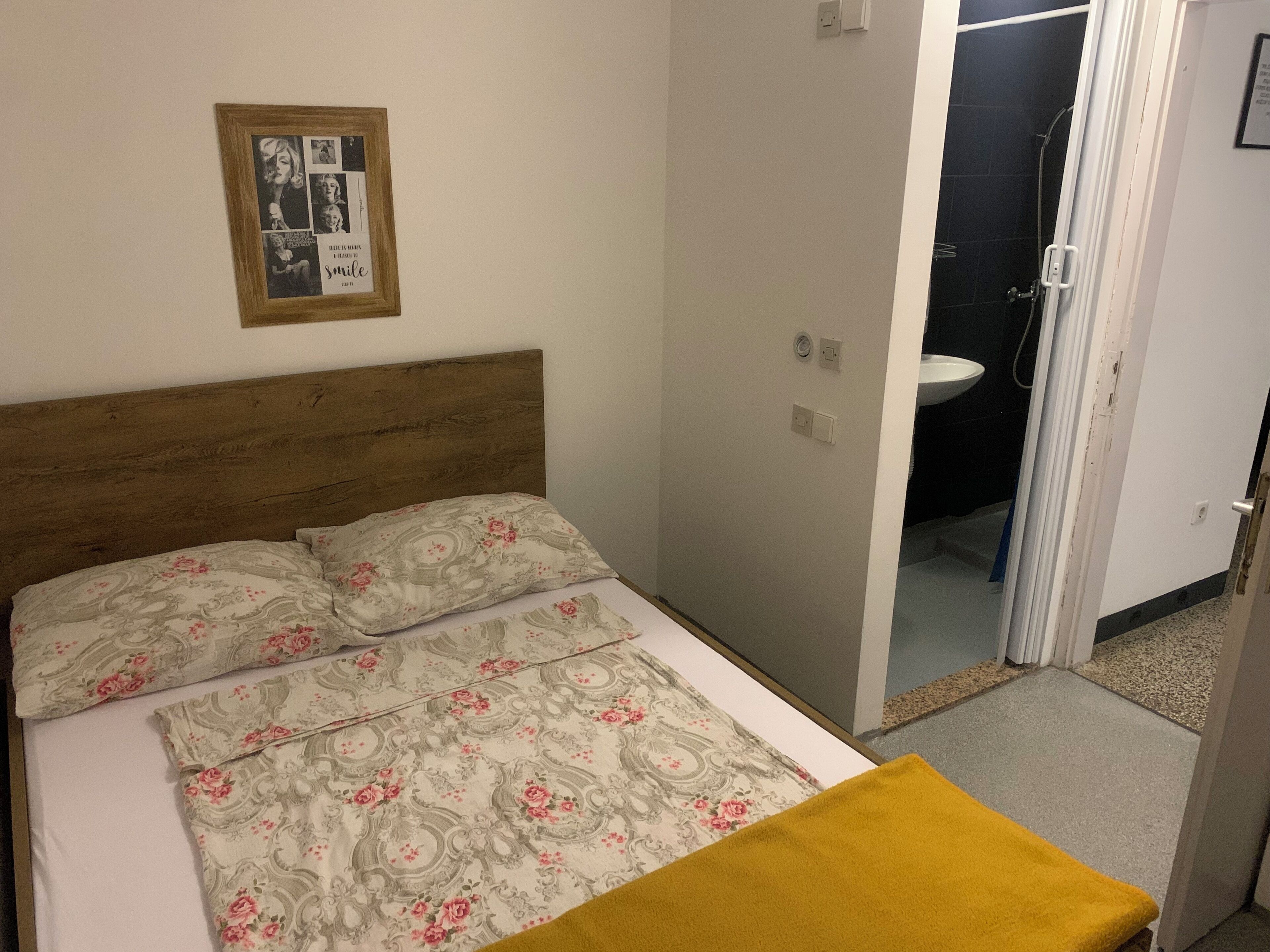 Un dormitorio con cabecera de madera, ropa de cama floral, un cuadro en la pared y un baño visible a través de una puerta abierta.