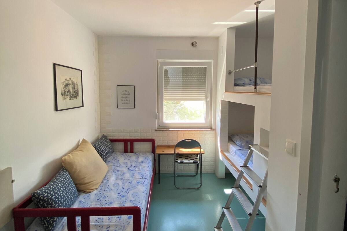 A small, narrow room with a single bed, a desk, and a chair. There is a window with blinds, a ladder, and a door.