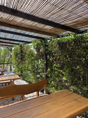 An outdoor patio area with wooden tables and benches, covered by a striped canopy, surrounded by lush greenery. - Hostel Kantun (Punat)