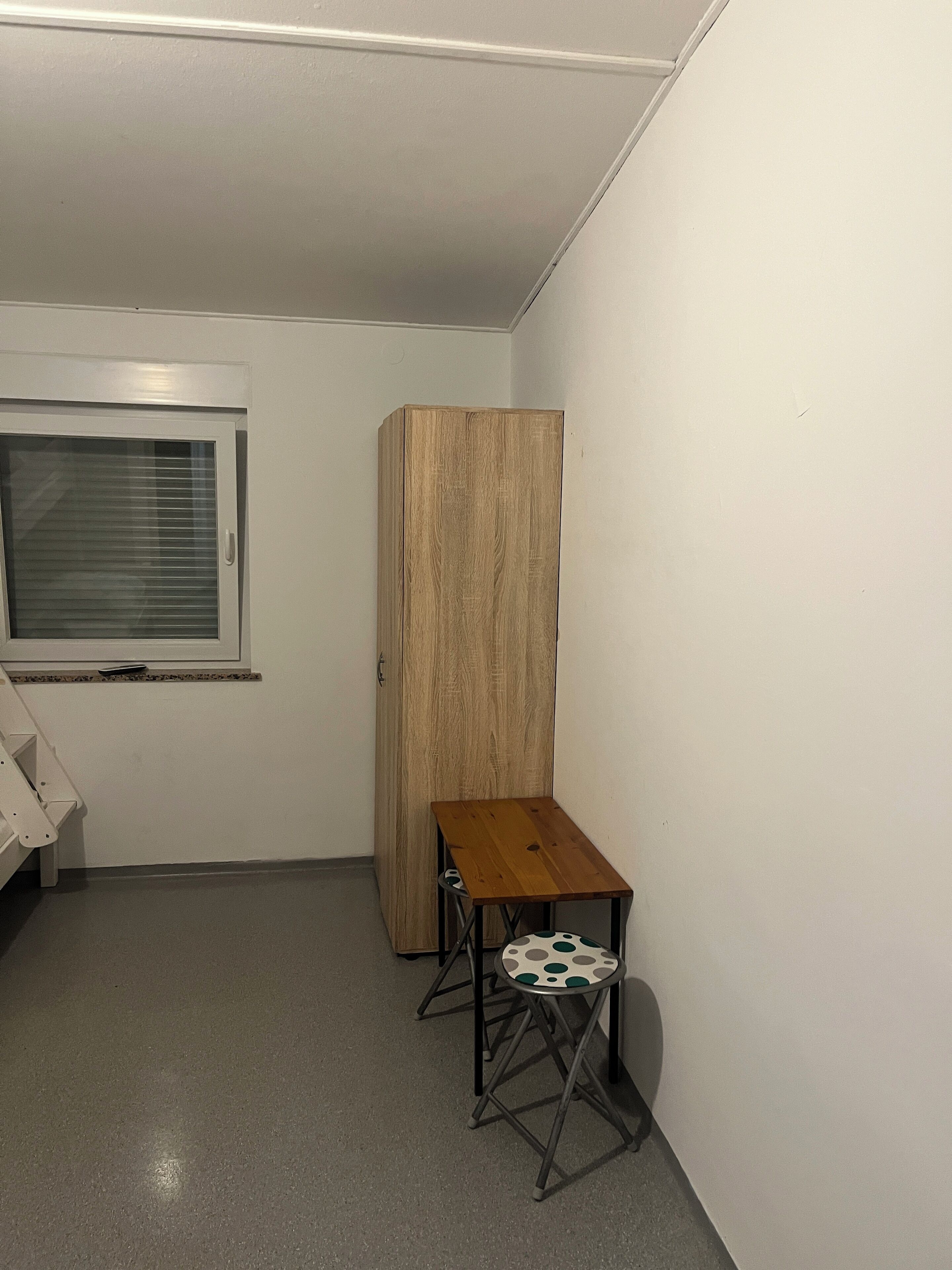 Una habitación pequeña y estrecha con un armario de madera, una mesa pequeña con un taburete y una ventana con persianas.