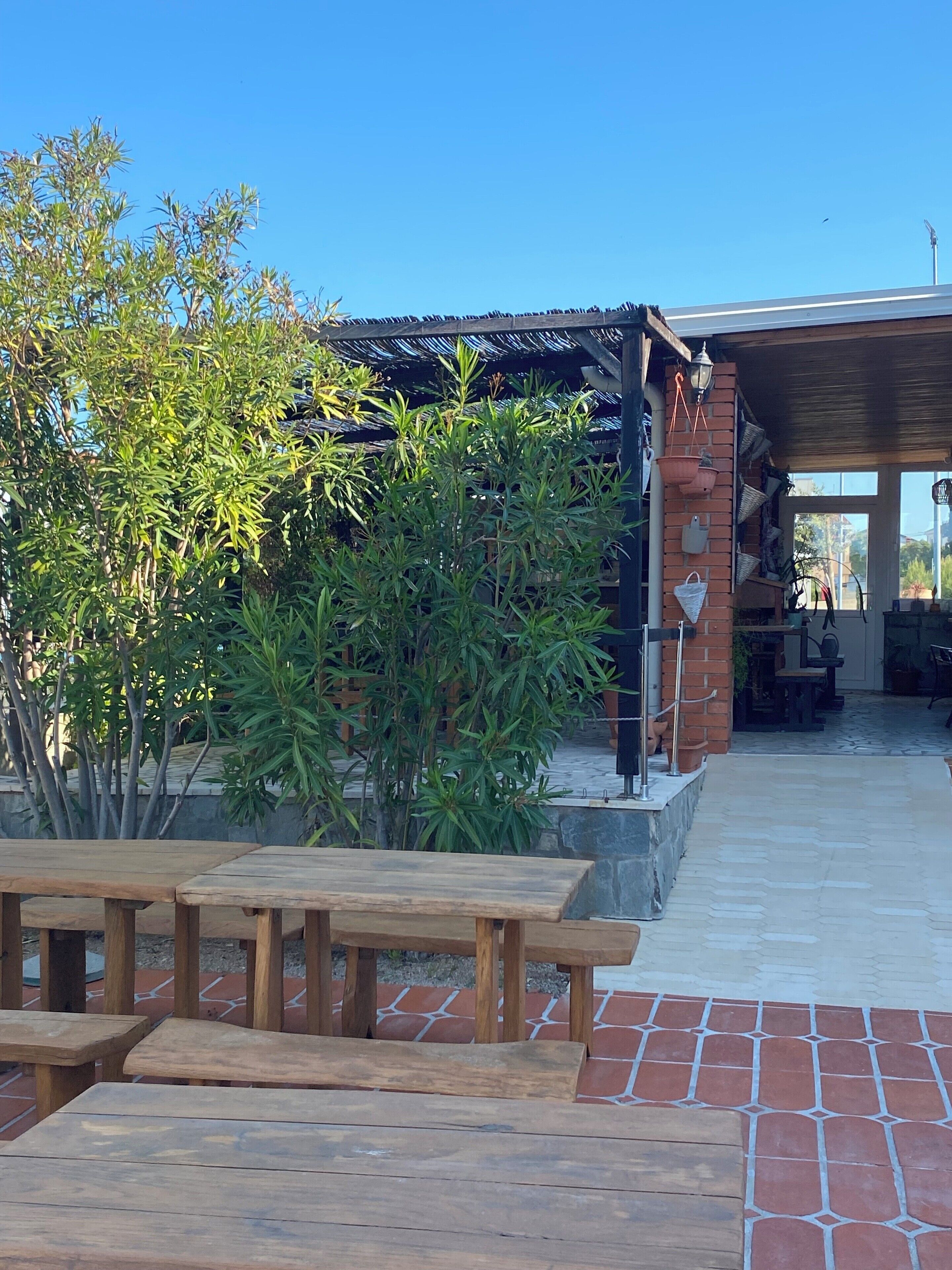 Un espace extérieur aménagé en terrasse, avec des tables et des bancs en bois, entouré de verdure et bordé d’un mur en briques.