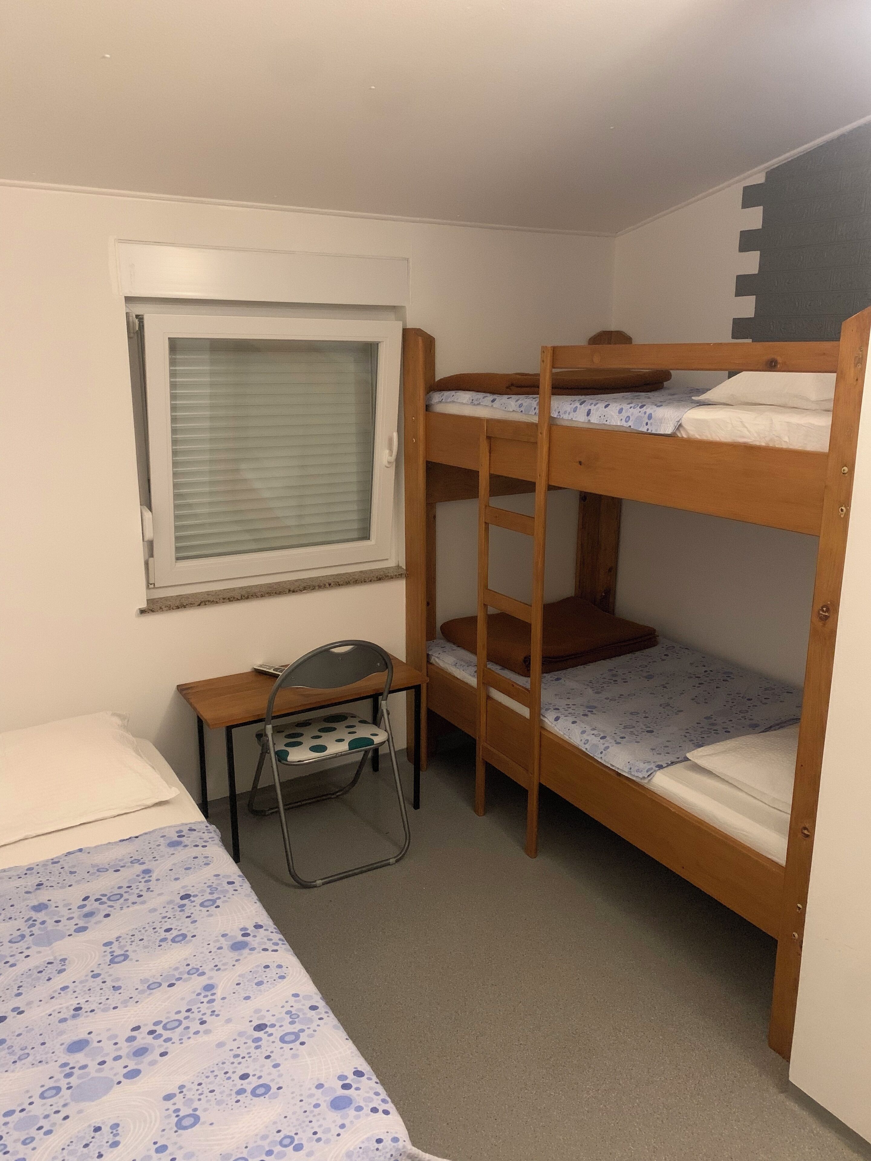 Habitación con literas, un escritorio y una silla, una ventana con persianas, y una cama con colcha estampada.