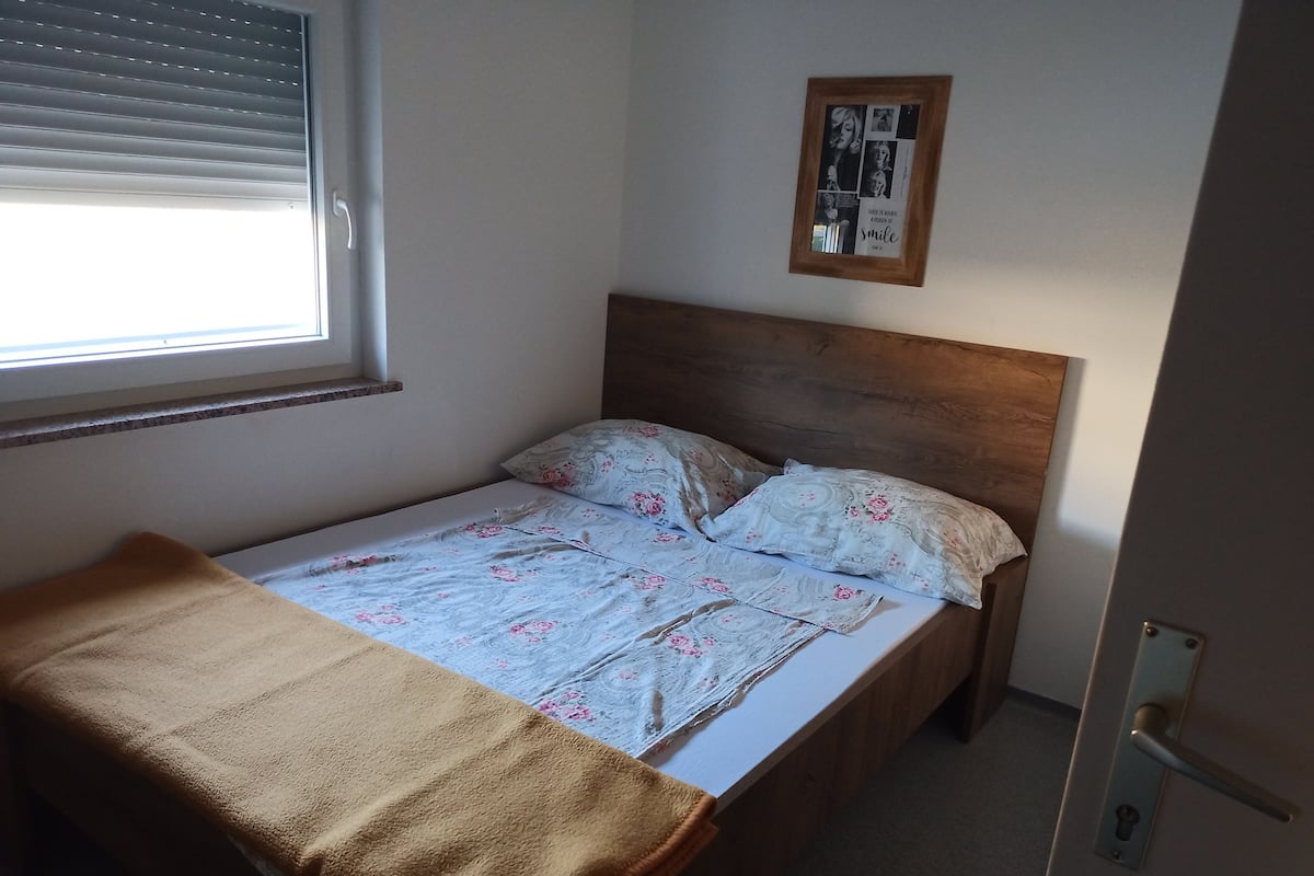 A single bed with a wooden headboard, a window with blinds, and a framed picture on the wall.