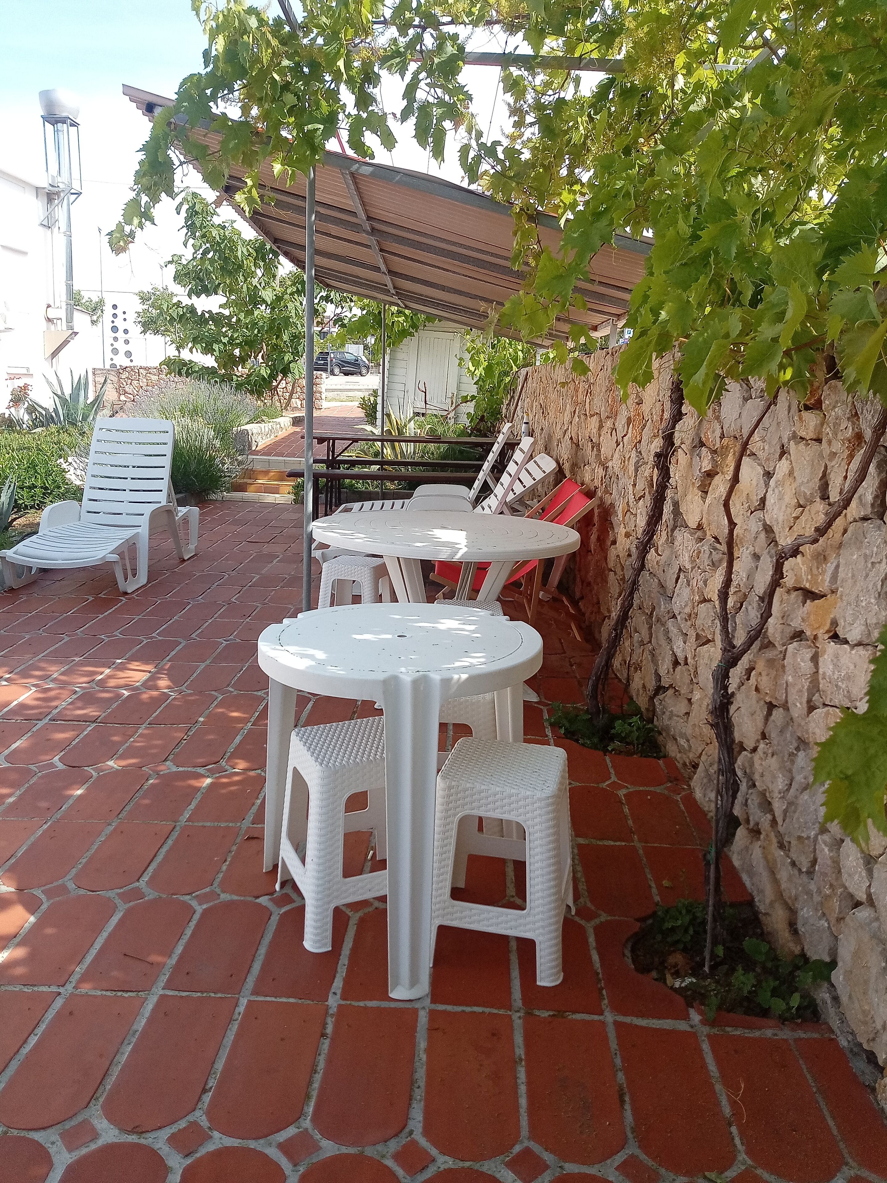 Un patio con muro de piedra, pérgola y mobiliario exterior, incluyendo sillas y mesas de plástico blanco.