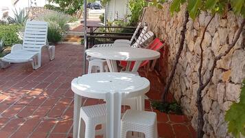 A patio area with a stone wall, a pergola, and outdoor furniture including white plastic chairs and tables.