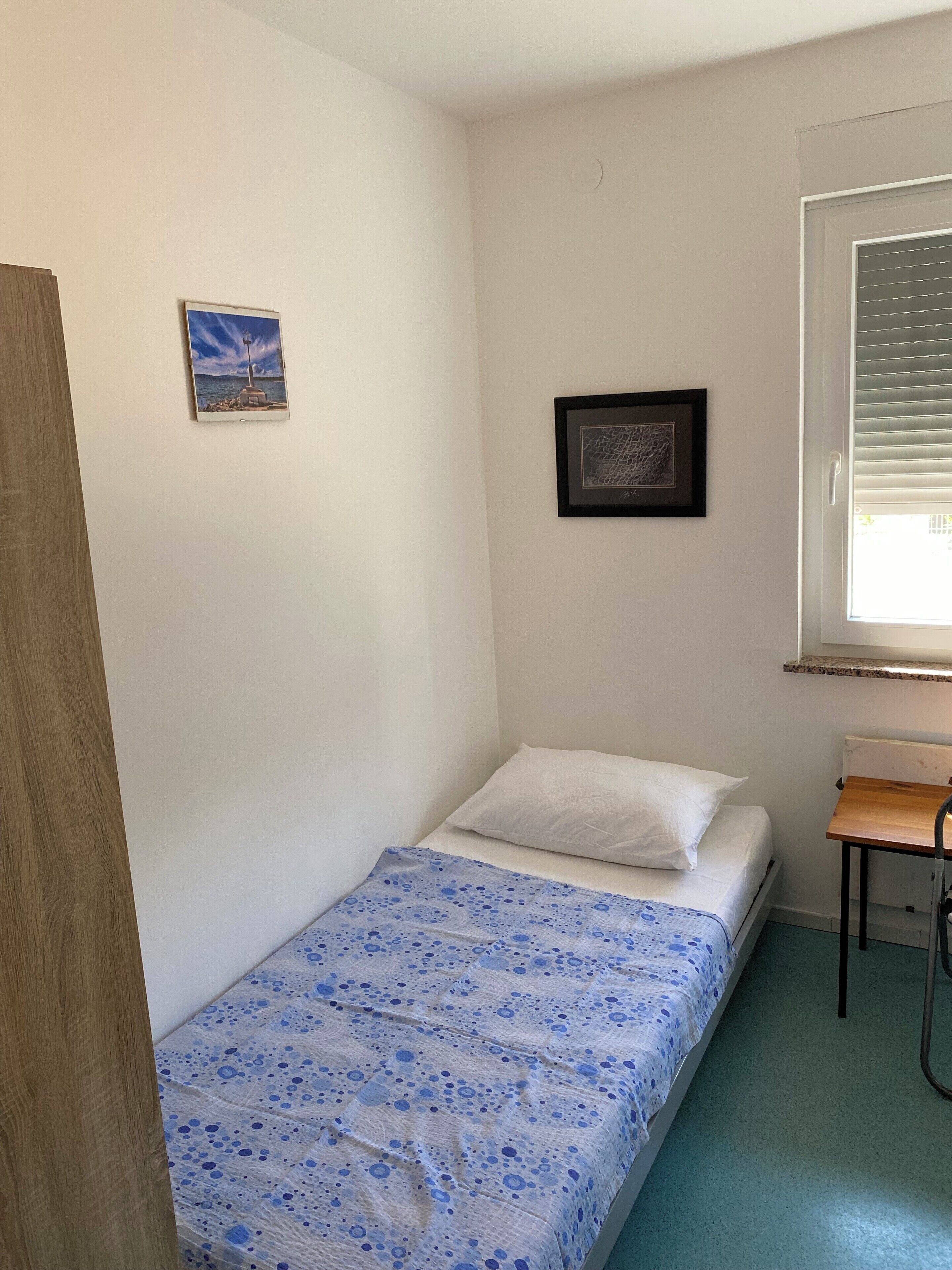 A small, single-bed room with a blue patterned bedspread, a white pillow, and a framed picture on the wall.