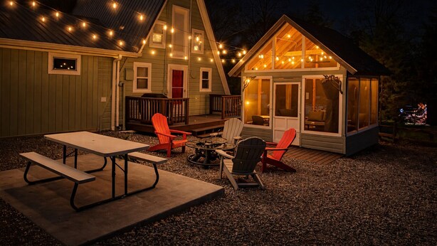 Un rincón acogedor al aire libre con una mesa de picnic y sillas Adirondack, una pequeña estructura de madera con puerta de vidrio y luces colgantes.