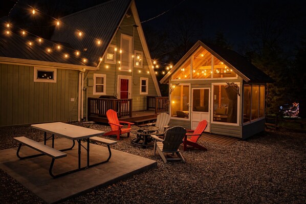A cozy outdoor seating area with a picnic table and Adirondack chairs, a small wooden structure with a glass door, and string lights.
