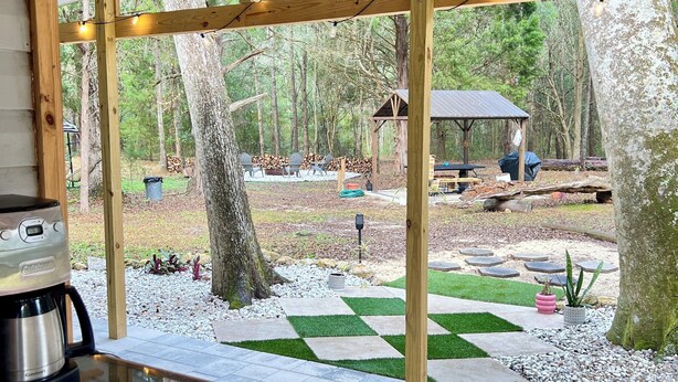 Un espacio al aire libre cubierto con una pérgola de madera, una cafetera sobre una mesa, y vistas a una zona boscosa con una barbacoa y asientos.