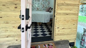Una cabaña de madera con piso a cuadros, una cama con papel tapiz floral y un felpudo que dice 'hello'.