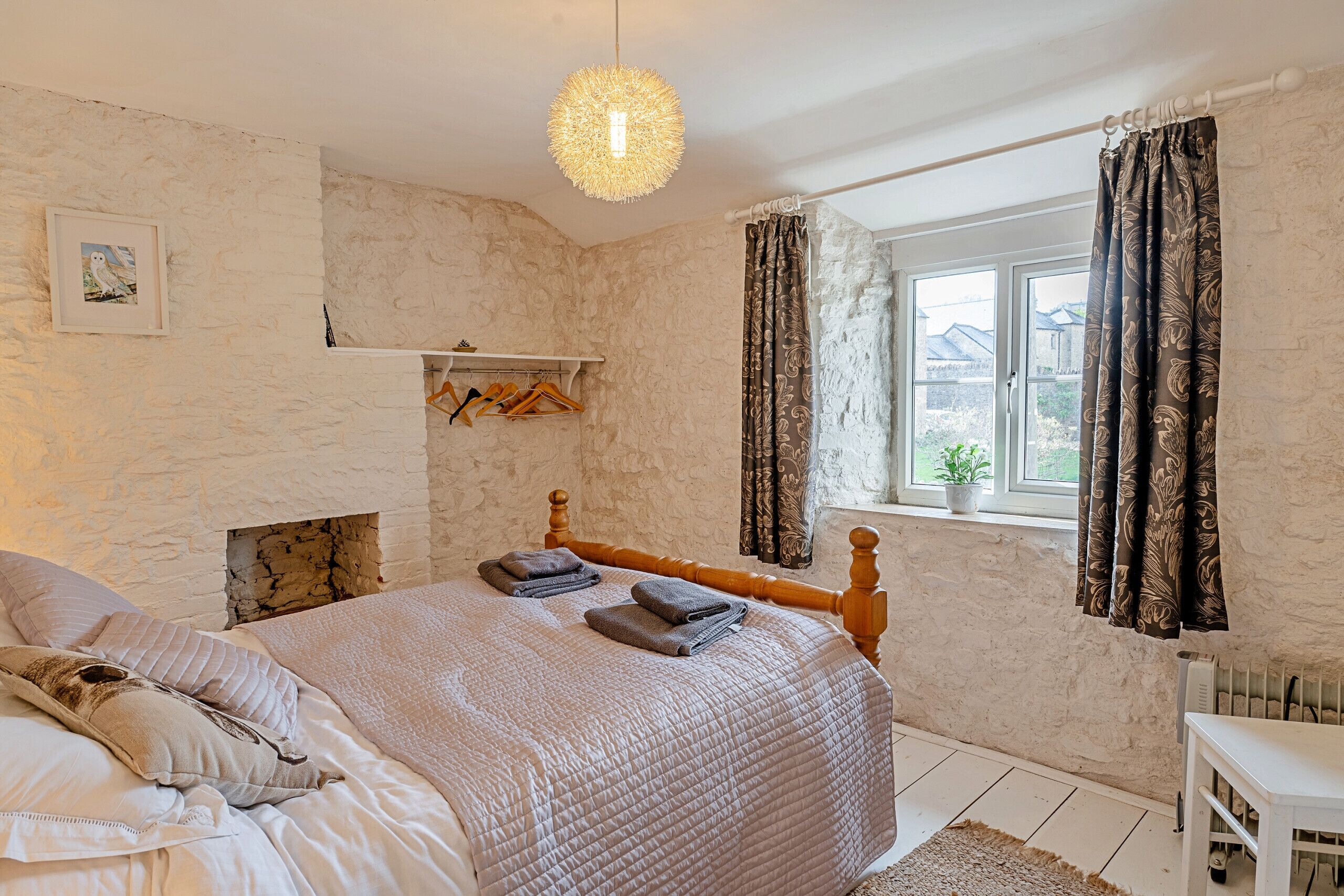 Ein Schlafzimmer mit Steinwand, einem Bett mit Holz-Kopfteil, einer Pendelleuchte, einem Fenster mit Vorhängen und einem kleinen Regal mit Kleiderbügeln.