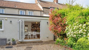 Una casa con patio, un giardino ricco di piante e un interno visibile con tavolo e sedie da pranzo.