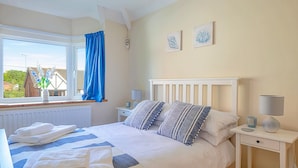 A neatly made bed with white and blue bedding, a bedside table with a lamp, and a window with blue curtains. - Sea Bank (Colwyn Bay)
