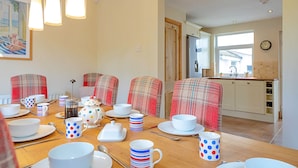 A dining area with a wooden table set for a meal, plaid chairs, and a kitchen in the background. - Sea Bank (Colwyn Bay)