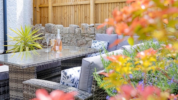 An outdoor seating area with a glass-top table, wicker chairs, and a stone wall backdrop. - Sea Bank (Colwyn Bay)